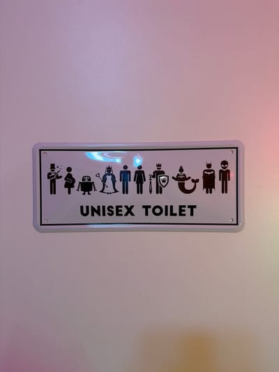 The image shows a vertical, rectangular, white unisex toilet sign with a grey border and black lettering/symbols. The symbols depict different figures stacked on top of each other. From top to bottom, they include an alien, Batman, a mermaid with a crown, a knight with a sword and shield, a standard male figure, a standard female figure, a penguin, a robot, and a magician. To the right of these symbols, the words "UNISEX TOILET" are vertically aligned. The sign is mounted on a white wall with a subtle gradient of pink and light yellow lighting. The location is implied to be in the city of Tarquinia, Italy, however, this information does not appear in the image itself.