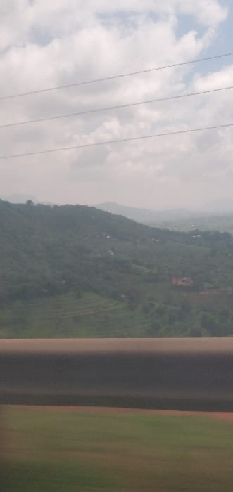 The image captures a daytime landscape in Kola ward, Kenya, framed from behind a dark gray, rounded horizontal barrier spanning the lower portion of the frame. The upper section of the image shows a sky with scattered white and gray cumulus clouds. Three thin, parallel utility lines are visible horizontally across the upper-middle sky. The middle ground features a succession of green, densely vegetated hills and mountains, which recede into the distance under atmospheric haze. Several small, distant structures with light-colored or reddish roofs are sparsely distributed across the hillsides, indicating human habitation. Below the foreground barrier, a blurred strip of green and reddish-brown ground is visible. No individuals are present in the scene.