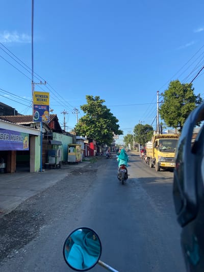 This is a street-level, first-person perspective photograph taken in the city of Margoyoso, Indonesia. The scene depicts a street with businesses lining the left side, including a storefront with a purple banner reading "SUMBER MURNI." A yellow sign above it also reads "SUMBER" and "MUSNI." Further down the street, other small businesses and street vendors are visible.
The road itself is paved and appears to be moderately trafficked. In the foreground, a motorcycle's rearview mirror is prominently visible, reflecting a person wearing a blue head covering. Ahead on the road, a person on a scooter, also wearing a blue head covering, is riding away from the camera. Further up the road, a yellow truck is traveling in the same direction.
The sky is clear and blue, indicating a sunny day. Trees line the right side of the street, and utility poles with numerous wires are scattered throughout the scene. The overall impression is that of a typical, busy street in a small Indonesian town.