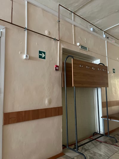 The image shows an interior hallway with light-colored walls and a textured ceiling. Copper pipes are run along the ceiling and down the walls, secured by metal clips. A white electrical outlet is mounted on the left wall, and a green exit sign with a right-pointing arrow is also visible on the wall. A red fire alarm pull station is mounted to the right of the exit sign. A wooden coat rack with multiple hooks stands in front of a doorway. The floor is tiled with a marble-like pattern, and red tape is visible on the floor. A wooden wainscoting runs along the lower portion of the left wall. This scene is likely within a public building in the city of Ikryanoye, Russia, possibly a school, administrative building, or other institutional facility given the utilitarian nature of the interior and the safety signage.