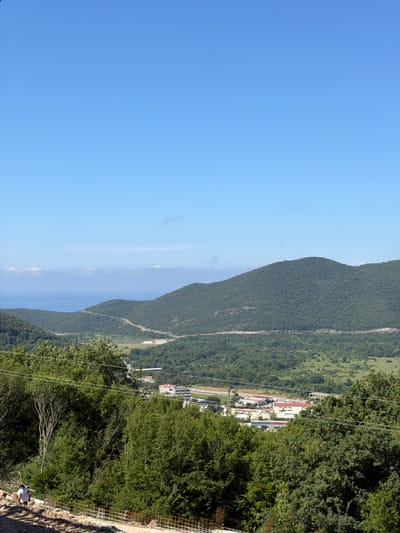 The image depicts a landscape scene, likely in Svinjišta, Montenegro, characterized by lush green hills and a clear blue sky. In the foreground, a person wearing a white shirt and a hat is visible on a dirt slope, seemingly engaged in some activity. Construction materials, including rebar, are present on the slope. A cluster of buildings, some with red roofs, can be seen in the middle ground, situated within a valley between the hills. A winding road is also visible, traversing the landscape. In the background, a body of water, possibly the sea, is visible beyond the hills. The overall impression is one of a rural or semi-rural environment with evidence of development.