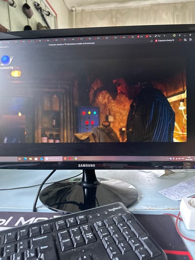 The image shows a computer monitor displaying a video streaming service with the Kinopoisk logo visible. The video content appears to be a scene from a movie or TV show, featuring a person wearing a striped shirt in a dimly lit, indoor environment. The computer's taskbar is visible at the bottom of the screen, showing the time as 15:03 and the date as 05.07.2026. A black keyboard with the "logitech" brand name is placed in front of the monitor. The background suggests an indoor setting, possibly a room with a desk or table, and some objects on a shelf are partially visible at the top of the image. The scene does not provide any specific indicators or context related to the city of Sinegorye, Russia.