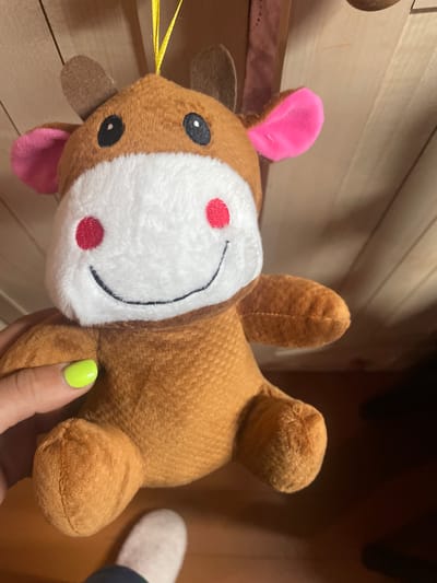 This is a close-up, eye-level, horizontal shot of a stuffed toy. The toy is lying on its back and appears to be a brown and white deer with pink ears and a pink tongue sticking out. It has black oval eyes and two red circles on its white muzzle, resembling cheeks. There is a black stitched smile. A yellow string is visible extending from the left side of the toy's head. A human hand with bright green nail polish is holding the toy's body. The background consists of a wooden wall, suggesting an indoor setting. The lighting is soft and appears to be from artificial sources. No text is visible in the image. The setting appears to be in a home environment in Sarzhenka, Russia.