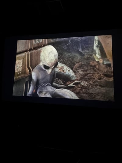 The image displays a screen, likely a cinema projection, showing a scene with an alien-like figure. The figure is humanoid in shape with a large, pale head, dark, oversized eyes, and thin limbs. Its skin appears to be a grayish-blue hue and has a textured, scale-like appearance. The alien is depicted in a seated position, leaning against a large, weathered, rectangular object that appears to be made of metal or a similar material. This object has visible seams and what might be labels or markings on its surface. The background is composed of rough, textured ground, resembling dirt and rocks, with some reddish-brown stains on some of the rocks. Other indistinct shapes are visible in the background, suggesting a larger environment, possibly a desolate or alien landscape. The lighting in the scene is dim, casting shadows and highlighting the textures of the alien and its surroundings. The overall scene appears to be from a science fiction film or media. There is no discernible location in Ondara, Spain, as the scene is fictional.