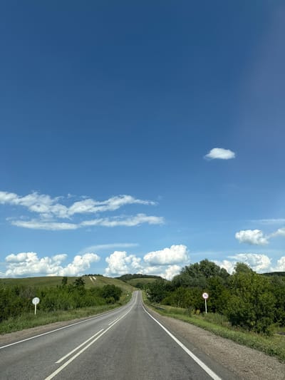 The image shows a road leading through a rural landscape in Chubovka, Russia. The road is asphalt with double white lines in the center and a white line on the right edge. To the left of the road, there is a round white sign with a blue circle and a diagonal line. Green grass and bushes line the side of the road, with more dense trees further back. In the background, there are rolling green hills with some trees and what appear to be tracks or paths on one of the hills. The sky is bright blue with scattered white clouds. A red and white circular speed limit sign with "70" on it is visible on the right side of the road. There are no people or animals visible in the image. The environment is natural and appears to be a sunny day.