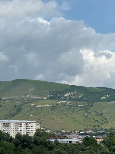 The image shows a landscape in Ust-Dzheguta, Russia, featuring a large, multi-story apartment building in the foreground, with a dense line of trees in front of it. Beyond the apartment building, a town with various residential houses and some commercial structures is visible. In the background, rolling green hills with sparse trees and some patches of exposed earth rise up towards a cloudy sky. The sky is partly blue with significant cloud cover, suggesting a mixed weather condition.