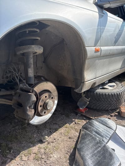 The scene depicts a silver-colored vehicle undergoing maintenance outdoors in Asenovo, Bulgaria. The front left wheel of the vehicle has been removed, exposing the brake rotor, caliper, and suspension assembly, which includes a spring and strut. The vehicle is elevated by a red hydraulic jack positioned beneath the frame, resting on a wooden plank, with a black support component atop the jack. Directly beneath the side of the vehicle, a removed wheel with a winter tire rests on the ground. The ground surface is composed of dry, uneven dirt with sparse patches of dry grass and scattered small rocks. In the foreground, a folded piece of reflective material, possibly a car sunshade or insulation, lies on the ground. A single red brick is visible near the removed tire. The scene is illuminated by strong sunlight, casting distinct shadows on the ground, indicating clear weather. No individuals are visible within the frame.
