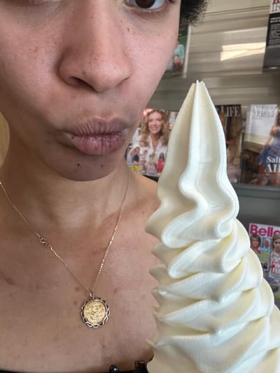 A close-up, vertically oriented, medium shot captures the left side of a person's face, focusing on their nose and lips, along with a portion of their neck and chest adorned with a gold necklace. The person, with curly dark hair partially visible on the left, appears to be looking away from the camera, with their lips slightly parted and glossy. The skin shows some natural texture and a few freckles.
Dominating the upper portion of the frame is a large, swirled soft-serve ice cream cone, its creamy white texture catching the light. The background is blurred, but suggests an indoor environment, possibly a store or market, with shelves of magazines visible. The magazine titles are partially obscured, but "LIFE" and "MAY" are discernible on some. Another magazine has "Belle" printed on it.
The lighting is bright, suggesting daytime. The scene seems casual, with the individual enjoying the ice cream, possibly as a treat or snack. The overall impression is one of a candid, everyday moment. There are no overt emotional expressions, but the focus on the food suggests enjoyment. The context of Westport, Ireland, is provided, though no specific landmarks are visible within the frame.