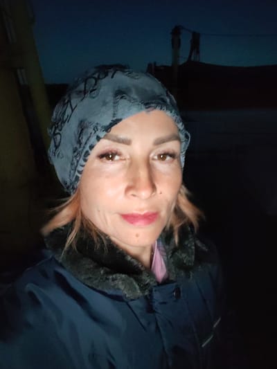 A woman with light brown hair and light skin poses for a selfie in front of a dark, outdoor background. She is wearing a blue jacket with a fur-lined collar and a blue patterned headscarf. Her eyes are brown, and she is wearing makeup, including eyeliner and lipstick. The background appears to be twilight, with a dark sky and dimly lit structures in the distance, suggesting a rural or suburban setting. It is not possible to determine the exact location within the city of Irdomatka, Russia from the provided image.