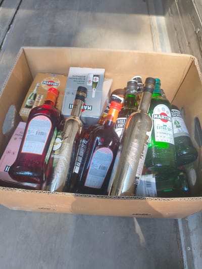 This image shows a cardboard box filled with various bottles of alcohol. The box is placed on a gray, rough surface that appears to be outdoors, possibly a pavement or concrete floor. Sunlight is casting shadows across the scene, suggesting it is daytime.

Among the bottles are several brands, including Martini. One bottle is labeled "MARTINI EXTRA DRY VERMOUTH." There are also bottles of what appears to be whiskey, with labels like "WILLIAM LAWS BLENDED SCOTCH WHISKY." Some bottles contain red liquid, while others contain clear or green liquid. The labels are primarily in English and Russian.

The overall impression is that of a collection of alcoholic beverages packed together, likely for transport or storage. The setting seems to be an outdoor, possibly industrial or urban area, given the concrete surface and the wooden planks visible in the background. There are no people or animals in the frame. The focus is solely on the contents of the box.