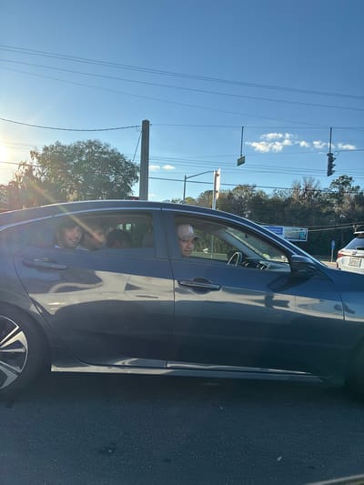 An outdoor, eye-level photograph depicts a dark blue four-door sedan or hatchback stopped in traffic on a sunny day in Gainesville, Florida, United States, specifically at the intersection of SW 34th Street and SW 20th Avenue.
Inside the vehicle, a male driver, wearing a light-colored baseball cap with a dark "NY" logo, is visible from the chest up, looking forward through the windshield. Behind him, in the left rear passenger seat, another male with dark hair is visible, smiling and looking slightly towards the left of the frame. A third individual, likely a child, is partially visible in the middle rear seat, with only the top of their head showing, looking forward. The back of a fourth person's head is discernible in the right rear passenger seat.
The background shows utility poles and power lines crossing a clear blue sky with sparse clouds. Bright sunlight originates from the left, casting strong light and reflections. To the far left, a red sign with the word "TACO" is partially visible. Further in the background, an intersection is evident with a hanging street sign displaying "SW 34th ST" and "SW 20th AVE," and a traffic light signal. A white SUV with a Florida license plate (partial: "FLS 05Y") is stopped directly behind the described sedan in the right background, indicating a queue of vehicles. Trees with varying foliage are present in the mid-ground. The road surface in the foreground is dark asphalt.