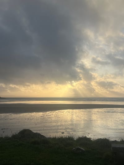 A beach scene unfolds with a golden glow. The sky is filled with dark and light clouds, the sun peeking through in the center. The beach extends into the distance, reflecting the golden light. Grass and some rock formations line the edge of the beach on the right side of the frame.
