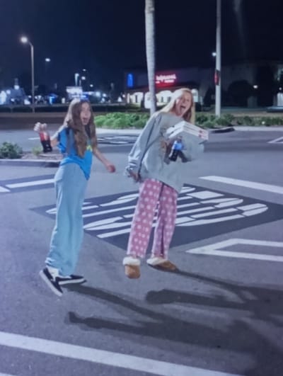 The image depicts two young females in mid-air over a paved surface at night, located in the city of Kukcha, Uzbekistan. The female on the left, with long light brown hair, wears a light blue t-shirt, light blue pants, and black and white athletic shoes. She holds a dark-colored bottle with a red label, identifiable as Coca-Cola, in her raised left hand, and her mouth is open in a wide expression. The female on the right, with long blonde hair, wears an oversized light grey sweatshirt, pink polka-dotted pants, and light brown fuzzy slippers. She carries several stacked white pizza boxes with red markings in her left arm, and her mouth is also open in a wide expression. Both individuals are positioned as if jumping, with their feet off the ground and casting elongated shadows on the dark grey pavement. The pavement features white painted markings, including a rectangular design with internal horizontal lines. In the blurred background, artificial lighting illuminates distant buildings, one with a red facade, along with streetlights and palm trees.