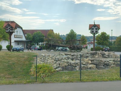 This is a wide, eye-level shot of the exterior of a restaurant in Himmelkron, Germany, with a large, antique horse-drawn wagon on display in the foreground. The restaurant building is white with a red-tiled roof and has a sign that reads "FRANKENFARM" above the entrance. There is a tall, decorative birdhouse on a pole to the left of the building, and another similar birdhouse structure to the right. In front of the wagon is a rocky landscape.
Several cars are parked in the background, including a blue hatchback, a dark sedan, and a black SUV. Two people are visible near the dark sedan, appearing to be loading or unloading items. A wire fence runs across the lower portion of the image, separating the viewer from the restaurant and display area. The sky is partly cloudy. The overall scene suggests a rural or semi-rural setting, likely a tourist attraction or roadside eatery.