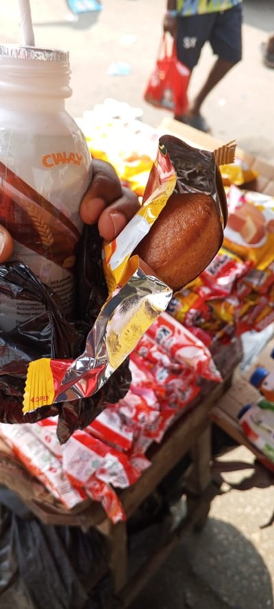A close-up photograph taken in Awjaw, Nigeria, depicts a dark-skinned hand holding two items. The left hand grasps a white plastic bottle, labeled "CWAY" with wheat graphics, containing a beverage, and fitted with a white drinking straw. The hand simultaneously holds a partially opened snack wrapper, exposing an oval-shaped, light brown pastry. The wrapper is primarily yellow and silver foil with red accents, showing "OPEN" on a red tab and "DATE: 216" visible. Beneath the items, a dark, crumpled plastic bag is present. In the midground, multiple individually packaged snacks, predominantly in red and white wrappers, are displayed on a wooden vendor's stall or table. The blurred background shows a person walking from left to right, wearing dark shorts and a colorful patterned shirt, carrying a red plastic shopping bag. The ground surface is light-colored, appearing to be dirt or concrete, with scattered debris. The scene is under bright daylight.