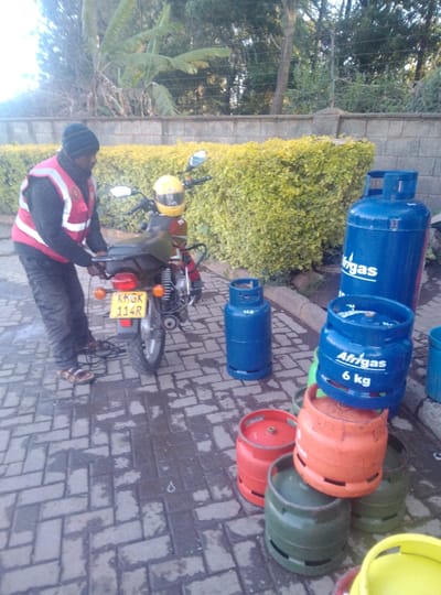 In Karai Ward, Kenya, a male individual is depicted standing on an interlocking brick pavement next to a red and black motorcycle. The man wears a black beanie, a dark long-sleeved garment, a red and white reflective vest, dark trousers, and sandals. He is positioned near the rear of the motorcycle, appearing to manipulate an object or secure something. A yellow motorcycle helmet rests on the motorcycle's handlebars. The motorcycle's rear license plate displays "KKGK 114R".
Adjacent to the motorcycle, a collection of approximately ten to twelve Liquefied Petroleum Gas (LPG) cylinders are arranged on the pavement. These cylinders vary in size and color, including blue, red, orange, green, and a partially visible yellow one. Prominently visible are several blue cylinders, one marked "Afrigas" and another specifically "Afrigas 6 kg". Other blue cylinders show a "12.5 kg" marking. The pavement exhibits some damp or wet patches.
Behind the subjects, a low, dense hedge with light green-yellow foliage runs along a concrete curb, separating the pavement from a planted area. Beyond the hedge, a light-colored stone or concrete wall is visible, backed by mature trees and lush green vegetation, including banana-like leaves. The scene is illuminated by natural daylight.
