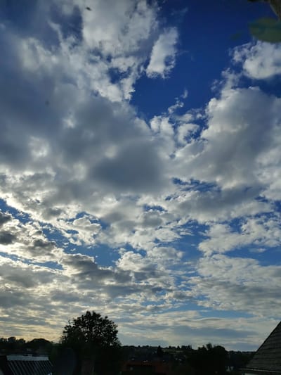 This media file is a vertical, landscape-oriented photograph taken from an elevated viewpoint, likely a balcony or window. The primary subject is the sky, filled with a dynamic array of clouds against a bright blue background. The clouds vary in density and color, with some appearing bright white and fluffy, illuminated by sunlight, while others are darker, suggesting depth and possibly approaching rain. Sunlight streaks through the clouds in places, creating highlights and shadows.
The lower right portion of the image shows a sliver of a built environment. A dark silhouette of a tree is prominent, with hints of buildings and rooftops beneath it. The scene suggests an outdoor setting during daylight hours, likely late afternoon or early evening given the quality of light and the presence of shadows on the landscape elements. There are no people or explicit objects of human activity visible in the sky portion, but the presence of buildings and a tree indicates a residential or semi-urban area. The weather appears to be fair but with considerable cloud cover. The overall mood conveyed is one of natural beauty and the changing patterns of the sky. There is no visible text in the image.
