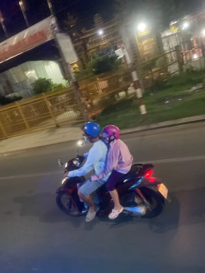 Two individuals, wearing helmets and masks, are riding a motorcycle at night on a paved road. The person in front, wearing a light blue long-sleeved shirt and shorts, is operating the motorcycle. The person behind, wearing a purple long-sleeved shirt and leggings, is a passenger. Both are wearing helmets, one blue and one pink. The motorcycle is dark-colored with a red taillight. The background shows a fence, greenery, streetlights, and buildings, suggesting an urban environment in Binh Thuan Ward, Vietnam. The overall scene is dimly lit, characteristic of nighttime.