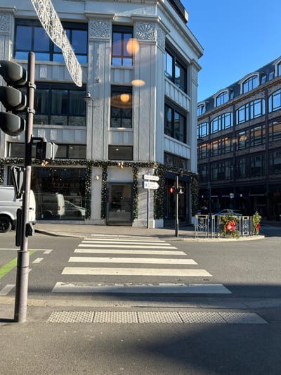 This image depicts a sunlit street intersection in Saint-Quentin, France, specifically at the corner of Rue d'Isle and Place du 8 Octobre. The scene captures a three-quarter view of an urban environment under a clear blue sky.
In the foreground, a pedestrian crosswalk with broad white stripes spans the asphalt road, bordered by tactile paving for the visually impaired. To the immediate left, a dark grey traffic light pole stands prominently, supporting a black traffic signal head and a pedestrian signal button. A faint green line, indicative of a bicycle lane, is visible on the street beside the pole.
Dominating the left and center-left is a multi-story building with a light-colored, possibly white or light grey, facade featuring Art Deco or early 20th-century architectural elements, including decorative fan motifs above some windows and vertical fluting. The ground floor displays large storefront windows, framed by green garlands adorned with small white lights, suggesting seasonal decoration. Reflections in these windows show interior lighting and a white panel van parked on the street, with the word "OUTLET" visible in reverse. Lens flares appear as orange circles in the upper windows of this building, indicating direct sunlight.
Across the intersection, on the right, stands another multi-story building with a darker facade, possibly brick or stone, characterized by numerous small-paned windows. Its ground floor also features large display windows. On the corner of this building, a low, decorative metal railing is decorated with festive evergreen branches and red accents. A red traffic light is visible further down the street to the right. One individual is partially visible standing on the sidewalk near the darker building.
Sunlight emanates from the left, casting long, distinct shadows across the crosswalk and onto the street. A white street sign on a pole is positioned near the corner of the light-colored building, indicating directions to "S. P. Vertes" (Sous-Préfecture / Place Verte) and "100 Places." No explicit actions or interactions are occurring among people or vehicles in the immediate captured moment, but the setting is active.