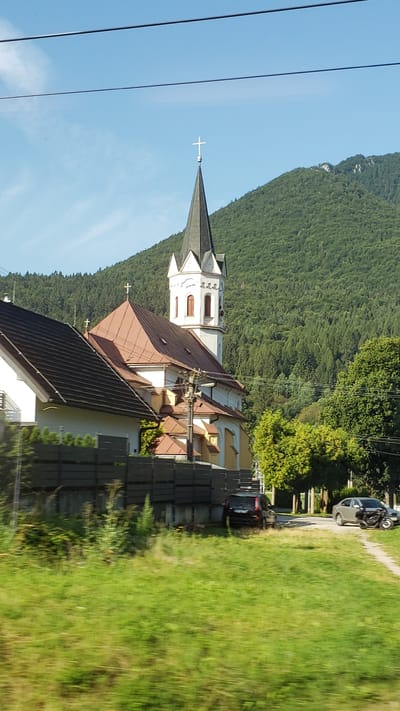 The image shows a church in Stankovany, Slovakia. The church has a tall, steeple with a cross on top. It is surrounded by trees and a mountain in the background. There are two cars parked in front of the church, one black SUV and one silver sedan. There is also a motorcycle parked next to the silver sedan. The sky is blue and there are some clouds. The image appears to have been taken from a moving vehicle, as the grass in the foreground is blurred.