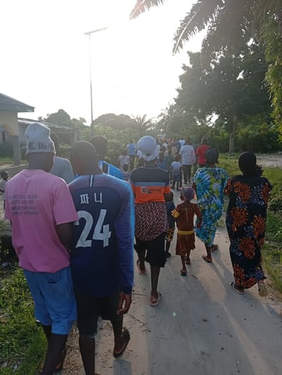A large group of people walks along a sandy road in Ibotirem, Nigeria, with houses and trees lining the path. The people, including adults and children, wear various casual clothes and sandals. A man in the foreground wears a pink shirt and blue shorts and walks next to a man with a blue shirt that has “24” on the back. The image is taken from behind the people. A light pole is visible.