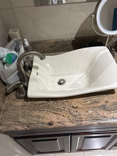 The image shows a bathroom sink with a white ceramic basin and a silver faucet. The sink is mounted on a granite countertop with brown, tan, and black streaks. There is a gray toiletry bag with toiletries on the counter to the left of the sink. There is also a small white square container and a small bottle of hand sanitizer on the counter. The image was taken from a slightly elevated position, looking down at the sink. The sink is located in a bathroom in a building at the coordinates 9.945, -84.201, which is in the city of Río Oro, Costa Rica. There is a mirrored makeup stand to the right of the sink. The lower part of the image shows a dark brown cabinet with two drawers. There is a portion of beige tile floor visible at the bottom of the image.