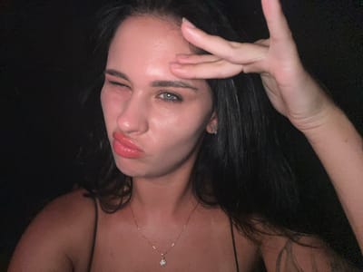 The image presents a close-up portrait of a female individual against an entirely dark, featureless background. She has long, dark hair. Her left eye is closed in a wink, while her lips are pursed into a prominent pout, accentuated by glossy coral-pink lipstick. Her right hand is raised, with fingers splayed and bent in a stylized gesture, touching her forehead. She is wearing a thin-strapped top, a small stud earring in her visible left earlobe, and a thin chain necklace with a small, clear gemstone pendant. A tattoo is partially visible on her right upper arm. Her skin exhibits a slight sheen under the direct lighting. No specific environmental details or location within Gangnam-gu, South Korea, are discernible from the dark background.