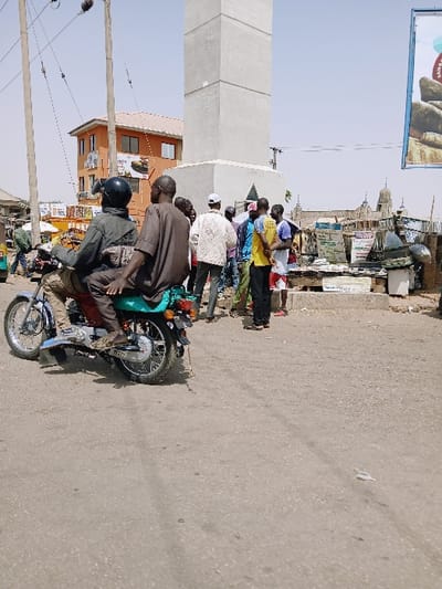 The image depicts a bustling urban street scene in Zambia, within the city of Kungulu School, under clear daylight conditions. In the immediate foreground, a grey asphalt road occupies the lower third of the frame. In the left midground, a blue and teal colored motorcycle is positioned, carrying two male occupants. The driver wears a black helmet, a dark long-sleeved shirt, and light-colored pants. The passenger, seated directly behind, wears a brown short-sleeved top and dark pants. Both individuals are facing away from the camera.
Further into the midground, on the right side, stands a tall, light-grey concrete tower or monument with a wider base. Adjacent to its base, a group of approximately eight to ten men are gathered, standing and appearing to interact or observe their surroundings. They are dressed in various casual attire, including shirts, t-shirts, and pants. Behind this group and extending to the right, numerous makeshift market stalls or vendor setups are visible, displaying goods and featuring various signs with text and images.
In the background, to the left of the concrete tower, a multi-story orange building with rectangular windows is visible. Overhead, multiple utility poles with a network of wires crisscross the sky. On the far right edge of the frame, a large, colorful vertical billboard with graphics of what appear to be food items or drinks is partially visible. The overall environment suggests a dry, sunny climate with a mix of permanent structures and informal commercial activity.