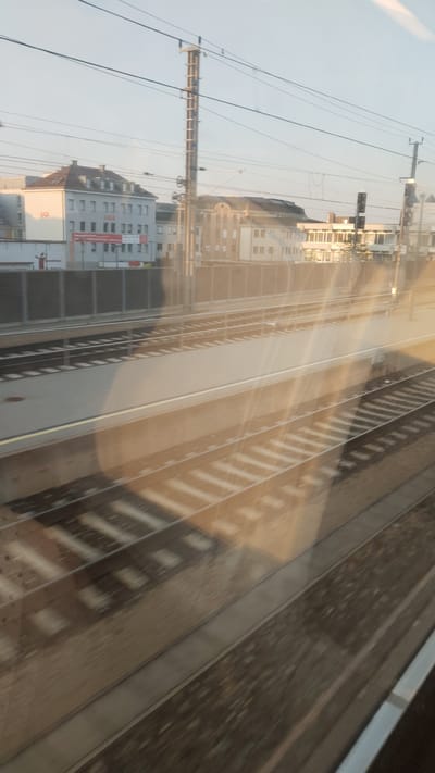 This media file captures a view from a train window looking out at a railway station and surrounding buildings. The scene depicts multiple train tracks, some with ballast and sleepers clearly visible. A platform area is also present, with a raised edge and a yellow line marking its boundary. In the background, several buildings are visible. One building on the left has "OGB" written in red letters and appears to be a multi-story structure with windows. Another building to its right is lighter in color and has an arched roofline. A tall utility pole with multiple wires stretches across the upper portion of the image. Sunlight is casting long shadows across the tracks, suggesting it is either early morning or late afternoon. The sky is a pale blue with hints of haze. The overall impression is one of a functional urban railway environment, likely in Sankt Pölten, Austria, given the context. There are no clear indications of specific activities or events taking place, nor are there any discernible emotions conveyed by the imagery. The presence of the train tracks and the perspective from within a moving train suggest the context of travel.