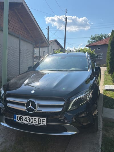 A black Mercedes-Benz car is parked on a paved surface in what appears to be a residential area. The car's front is prominently displayed, with its grille, headlights, and license plate visible. The license plate reads "OB 4305 BE" and below it, "BG" indicating it is from Bulgaria. The car has a Mercedes-Benz logo on its grille.

The setting suggests a sunny day, possibly late morning or early afternoon, given the bright sunlight and the presence of shadows. The sky is blue with scattered white clouds. In the background, there are houses with tiled roofs, some with wooden elements, and a chain-link fence. Utility poles and power lines are also visible, suggesting the presence of infrastructure. The overall scene depicts a quiet, everyday moment in a somewhat rural or suburban environment. There are no people visible in the image. The sunlight reflects off the car's surface, creating highlights and glare. The vegetation consists of trees and grass, contributing to a natural outdoor setting.