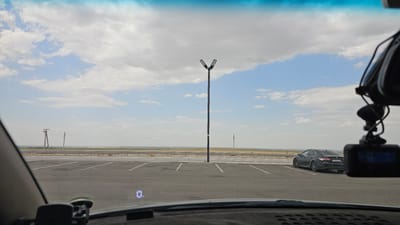 The image shows a wide, empty parking lot with asphalt paving and white parking space lines. In the distance, a highway is visible, separated from the parking lot by a metal guardrail. A dark-colored sedan is parked in the parking lot. A tall streetlight with two lamps stands in the center of the lot. Power lines and utility poles are visible in the background, stretching across the landscape. The sky is partly cloudy with a mix of blue sky and white clouds. The scene is likely located in an open, rural area. The image appears to be taken from inside a vehicle, with a dashboard and windshield visible in the foreground. A dashcam is mounted on the windshield. The location is identified as разъезд Кайнар, Kazakhstan.
