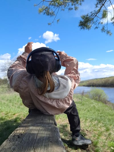 An individual with long brown hair, wearing a black baseball cap and black over-ear headphones, is captured in a natural outdoor setting. They are dressed in a light, muted pink or beige windbreaker jacket, black trousers, and black athletic shoes. The person is positioned on or leaning against a large, weathered wooden log or beam, with their body angled downwards, seemingly engrossed in an activity like listening to music or a podcast. Their right foot is visible, resting on the vibrant green grass of a riverbank.

The background features a calm river stretching horizontally, with a dense, green treeline visible on the distant bank. The sky above is a bright, clear blue, adorned with scattered white, fluffy clouds, indicating a sunny day. A pine tree branch with green needles extends into the frame from the upper left, adding to the natural ambiance. The overall scene suggests a peaceful moment of relaxation amidst nature, likely during late spring or summer. The location is Elets-Malanino, Russia. No visible text is present.