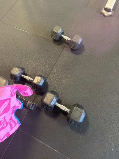 The scene is located in a gym in Neuville-aux-Bois, France, on a dark, textured rubber floor composed of interlocking mats. Three hexagonal dumbbells are present: one 20KG dumbbell positioned in the upper right, a 17.5KG dumbbell in the mid-left, and a 9.0KG dumbbell in the lower right. A pink and blue patterned towel lies partially visible in the lower-left quadrant, covering part of a black spray bottle with its nozzle pointed left. A segment of a metal base from gym equipment is visible in the upper right background. No individuals are present, and no actions or interactions beyond static placement are depicted.