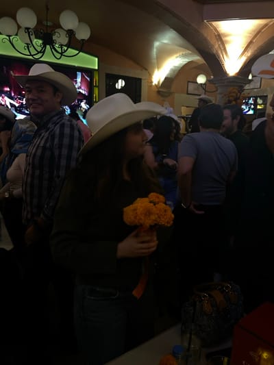 The image displays an indoor gathering occurring within a venue in Guanajuato City, Mexico, characterized by warm, dim lighting and historic architectural features including arches and stuccoed walls.

In the left foreground, a man wearing a light-colored cowboy hat and a plaid shirt looks towards the left with a slight smile. Directly to his right, in the center foreground, a woman with long dark hair, also wearing a white cowboy hat, stands in profile facing right. She holds a bouquet of vibrant orange flowers, consistent with marigolds (cempasúchil), tied with an orange ribbon. Her dark long-sleeved top and blue jeans are visible.

The midground contains multiple individuals. A man in a gray t-shirt stands with his back partially to the camera, appearing to engage in conversation with another man to his right. Other figures, some also wearing hats, are present in the crowded space.

The background reveals architectural details: a large black wrought-iron chandelier with five spherical white light fixtures hangs from the ceiling. Multiple arched passageways are visible, illuminated by upward-directed lights that highlight their contours. Two large screens display indistinct imagery; one is behind the man in the plaid shirt, and another is partially visible to the right. On a surface in the lower right foreground, a dark patterned handbag, a blue-capped bottle, and glassware are arranged. Additional orange flowers are present as decoration in the background, particularly beneath an illuminated arch.