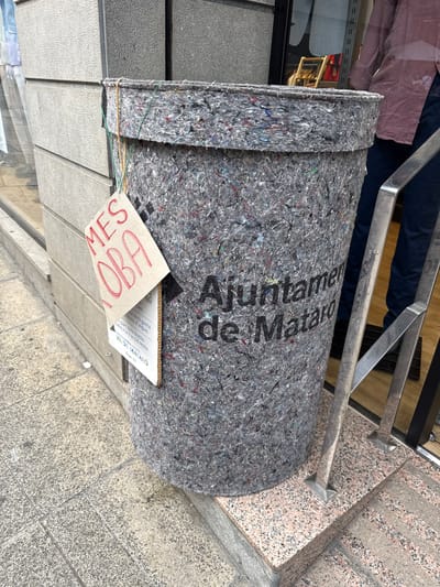 The image shows a large, cylindrical trash receptacle made of recycled materials, situated on a sidewalk in Mataró, Spain. The receptacle is gray and appears to be covered with a rough, felt-like material containing colorful flecks. A sign is attached to the side of the receptacle, written in red marker on a piece of cardboard. The sign has two lines of text: "MES" and "JOBA." Beneath this, a smaller, white sign with black text is partially visible, indicating it is related to textiles and the municipality. The receptacle is located next to a building facade with a light-colored, textured finish and a reflective window. A metal railing with a silver finish is also visible to the right of the receptacle, leading up some granite-clad steps. A person wearing a pink shirt and dark pants is partially visible in the background, standing behind the railing. The ground is paved with textured concrete slabs and granite.