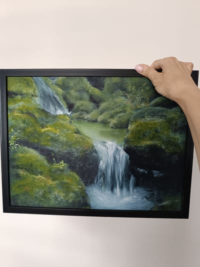 A hand with a pink nail is holding a framed painting of a waterfall. The painting depicts a lush green landscape with water cascading down moss-covered rocks into a pool. The artwork is framed in black. The background is a plain, light-colored wall. There is no indication of the location being Olsztyn, Poland.