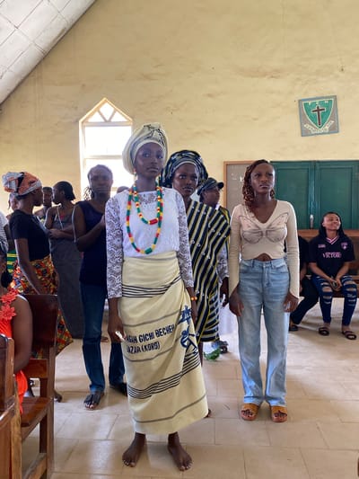 In the city of Dutsen Kato, Nigeria, a group of women stand in a room with a light-colored, textured wall and a sloped ceiling. The floor is tiled. In the center of the image, a woman in a white lace top and a cream-colored skirt with blue and black stripes stands barefoot, adorned with a colorful beaded necklace. To her left, a woman in a black t-shirt and a patterned skirt also stands barefoot. Behind them, other women are visible, some in traditional attire and head coverings, others in modern clothing like jeans. One woman wears a black t-shirt with the word "VICTORY" in pink. A green and white crest with a cross is mounted on the wall to the right. A doorway with green doors is visible in the background. The overall scene appears to be a gathering or event, possibly in a community hall or church.