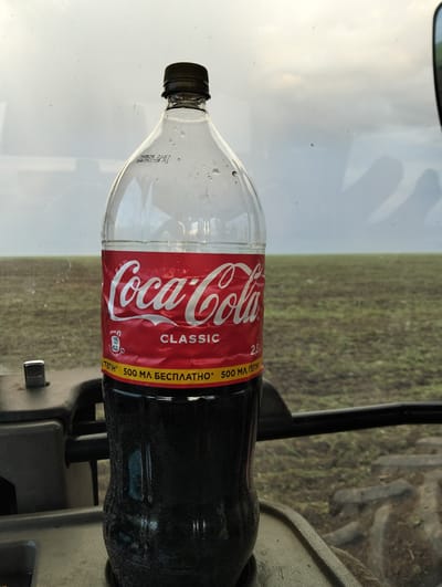 A Coca-Cola Classic bottle is positioned on the dashboard of a vehicle, likely a tractor, with a dark, carbonated liquid inside. The bottle's label is red with white lettering that reads "Coca-Cola" and "CLASSIC." Below this, text in Ukrainian indicates "500 ML FREE." The scene appears to be outdoors, with a blurry field and a cloudy sky visible through the vehicle's windshield. The location is identified as the city of Titov, Kazakhstan, suggesting the vehicle is likely in an agricultural setting. There are no people visible in the image. The overall lighting is subdued, consistent with overcast weather.