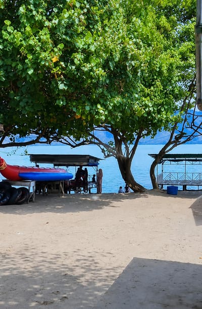 This is a photograph of a beach scene in Situngkir, Indonesia. The foreground is a sandy beach with shadows cast by trees. In the mid-ground, a bright blue body of water is visible, with a distant shoreline of mountains or hills. To the left, a red and blue inflatable boat is resting on a wooden platform alongside several black tires, presumably for water activities. Behind the inflatable boat is a covered structure with a corrugated roof, where several people are gathered. To the right, another covered structure with white railings stands near the water. Two individuals are seated near the water's edge in the right mid-ground, and a person in a light-colored headscarf and dress stands near the covered structure on the left. A large tree with lush green foliage dominates the left side of the frame, its branches extending over the beach and water.