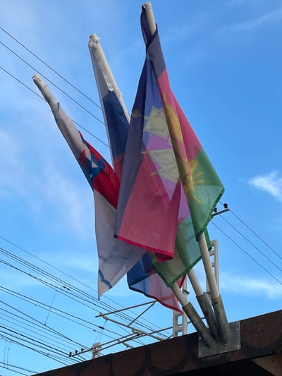 Three flags are mounted on poles attached to a dark, textured surface, likely the roof of a building. The flags are blowing in the wind against a bright blue sky with scattered white clouds. The leftmost flag is white with a red and blue emblem. The middle flag is white with a blue stripe and appears to be draped over the leftmost flag. The rightmost flag is a vibrant display of colors including pink, purple, green, and yellow, with a yellow emblem in the center. The flags are positioned against a backdrop of overhead electrical wires and a white metal structure. The image is taken from a low angle, looking upwards.