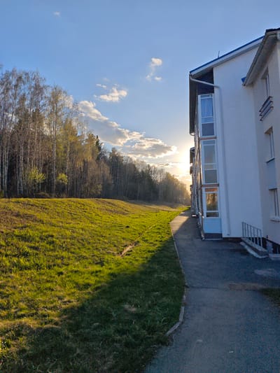 The image shows a view in Мичуринский, Russia, featuring a multi-story white building to the right, partially obscuring the view of the setting sun. The building has blue trim and several vertical window bays. A paved walkway runs along the side of the building. To the left of the walkway is a grassy embankment, and beyond that, a line of trees with some new spring foliage. The sky is blue with scattered clouds, illuminated by the sun. The light suggests it is either sunrise or sunset. No people are visible in the image.