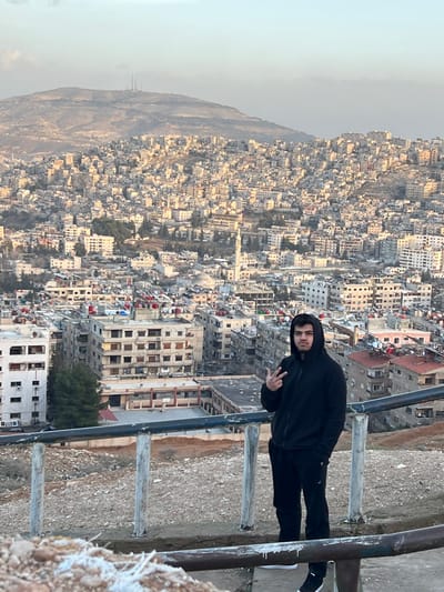 A young male stands in the foreground, facing slightly left while looking directly at the viewer. He is wearing a black hooded sweatshirt with the hood up, black track pants, and black athletic shoes with white soles. His right hand is raised, forming a "peace" sign (V-shape with index and middle fingers), while his left hand is in his pocket. He is positioned behind a metal railing, consisting of white vertical posts and horizontal bars, overlooking a expansive urban landscape.

The scene is an elevated panoramic view of the city of Qudsaiya, Syria. The city sprawls across undulating terrain, densely packed with numerous light-colored, multi-story residential buildings. A prominent white minaret of a mosque is visible within the city's mid-ground. Many rooftops feature red-capped water tanks and satellite dishes. In the distant background, a large, barren mountain range is visible, topped with several communication towers against a hazy, light-blue sky. The immediate foreground around the subject consists of loose dirt, rocks, and sparse dry vegetation.