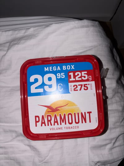The image displays a red and blue rectangular container for "PARAMOUNT VOLUME TOBACCO" which is placed on a white, textured surface that appears to be bedding. The container lid has prominent text indicating "MEGA BOX" and pricing information: "29 95 €" and "125g". Below this, "BIS ZU 275*" and "STÜCK" are visible, along with a small serial number "127360". A stylized red bird graphic is present above the "PARAMOUNT" branding. The lighting is dim, with shadows suggesting an indoor environment. There are no people or animals present in the image. No specific location in Münster, Germany, can be determined from the visual information provided.