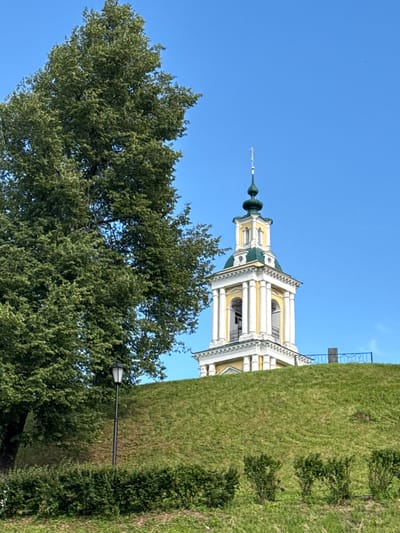 The image depicts the bell tower of the Resurrection Cathedral in the city of Vereya, Russia. The bell tower is white with yellow accents and a green roof, topped with a cross. It stands on a grassy hill, with a large green tree to its left. In the foreground, there is a street lamp and manicured bushes. The sky is a clear, bright blue.