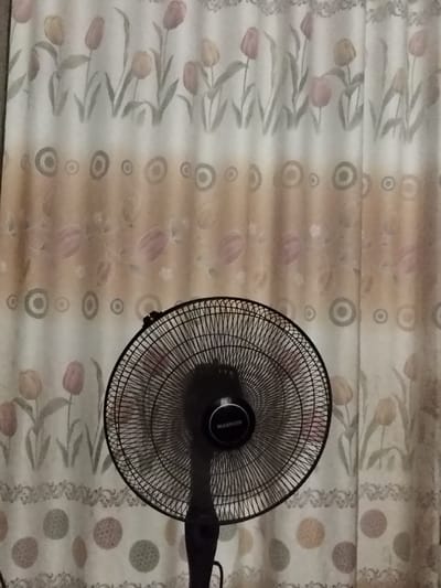 A black standing fan dominates the foreground, obscuring the curtain behind it. The curtain features horizontal bands of designs: a row of tulips, a line of circles, another tulip pattern, another line of circles, and another tulip pattern. The colors are muted, mostly tans, grays, and pinks. The lighting is soft, creating a slightly faded effect.