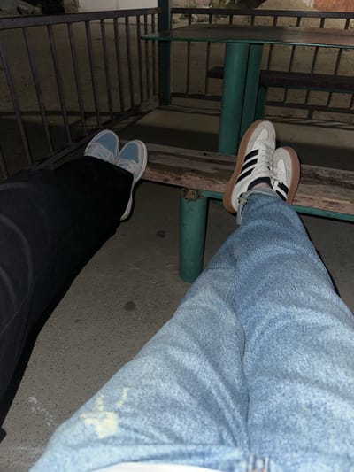 This low-light photograph captures the legs and feet of two individuals in an outdoor or semi-outdoor setting in Agarak, Armenia, likely at night.

One person, positioned towards the top of the frame and seen from the knees down, wears light blue, possibly acid-wash, jeans. Their feet are clad in white sneakers featuring three prominent black stripes on the side and a tan gum sole, indicative of an Adidas Samba style. These feet are casually propped on a weathered wooden beam, which appears to be part of a larger structure, adjacent to a dark green metal pipe support.

The second person, occupying the lower portion of the frame, also appears from the knees down. They are wearing black pants and light blue and white sneakers. Their feet rest on a rough concrete surface, next to a dark, open metal grate, which strongly suggests a staircase, platform, or utility access point.

The overall scene is characterized by industrial or functional architectural elements, including the concrete ground, metal grating, green pipes, and worn wooden planks. The dim lighting creates strong shadows and a somewhat muted color palette, emphasizing the texture of the materials. The postures of the individuals suggest they are casually seated or resting in this urban or industrial environment. There is no visible text in the image.