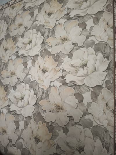 A close-up, head-on shot shows a wallpaper pattern featuring large, detailed floral designs with subtle hints of pale peach and cream on a muted gray background. The flowers are rendered with a sense of depth and texture, with visible stamens and layered petals. The image is a still life, devoid of any human presence or urban context within the city of Умыган, Russia.