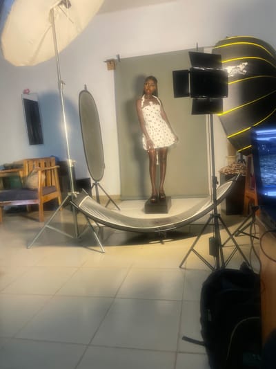 In what appears to be a photography studio in South Ropp, Nigeria, a young Black woman is posing for a photo shoot. She is wearing a short, white polka-dot dress with a halter neckline and black platform heels. She stands on a black rectangular pedestal.
The studio is equipped with various photographic lighting equipment. A large, white umbrella light is positioned to the left of the subject, supported by a silver stand. In front of the subject and slightly to the right is a curved reflector, also on a stand. To the right of the subject, a large softbox light with yellow accents is set up. Several smaller lights are attached to a stand in front of the softbox. A dark gray backdrop is behind the woman.
In the background, to the left, a wooden chair with blue cushions is visible. A mirror hangs on the wall behind it. In the right foreground, a black backpack rests on the tiled floor, and a monitor displaying an image is on a wooden surface. The floor is tiled with light-colored squares. The overall lighting suggests an indoor artificial lighting setup typical for a studio environment.