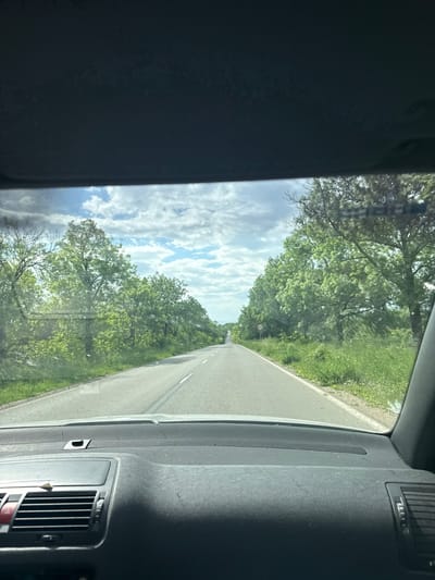 The view from inside a vehicle driving on a road. The road is straight and lined with green trees and vegetation. Above the trees, the sky is visible with scattered white clouds. The vehicle's dashboard and interior are visible on the sides of the frame. The location is possibly Studeno Buche, Bulgaria.