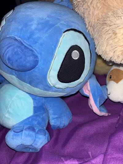 The image presents a close-up view of several plush toys, dominated by a blue plush toy of Stitch from Disney's Lilo & Stitch. Stitch is positioned prominently, with its large, black oval eye featuring a white embroidered pupil clearly visible. Its light blue facial features and belly stand out against its darker blue body. Pink inner ear linings are visible on one of Stitch's ears.

Partially obscuring Stitch's head on the upper left is a portion of a light brown, shaggy plush toy. Above Stitch's ear, another small, white plush toy with brown markings is visible.

The background consists of a deep purple fabric, which appears to be a blanket or bedding, suggesting an indoor setting such as a bedroom or living room. The fabric exhibits folds and a noticeable weave. The lighting is warm and somewhat dim, indicating an indoor environment, possibly during the evening or night.

No specific activity is occurring; the toys are simply arranged together. There are no discernible location cues specific to Cherniche, Bulgaria, beyond the general domestic setting. No text is visible in the media file. The overall impression is one of comfort and a personal, cozy space.